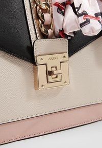 Beige and black structured handbag with gold hardware, featuring a chain accent and fabric scarf detail. Textured faux leather material.