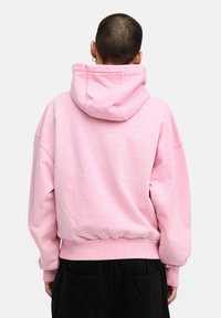 Pink hoodie with a rounded hem, oversized fit, and a large adjustable hood. Soft texture and ribbed cuffs. Back view.