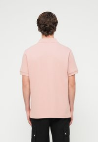 Polo rose clair, manches courtes, avec col. En coton, présente une texture lisse et des détails de couture minimaux.