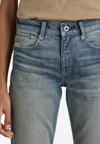 Jeans de mezclilla azul claro con un ajuste slim, que cuentan con un diseño de cinco bolsillos, costuras visibles y una pequeña etiqueta con el logo en el bolsillo delantero.