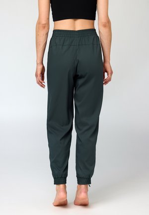 Person iført mørkegrønne joggebukser med elastisk talje og sort crop top, stående barfodet med ryggen vendt mod en ensfarvet lys baggrund.