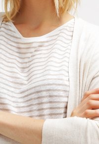 Ljusbeige topp med horisontella, tunna vita ränder, med rund halsringning och mjuk textur, lagd under en ljus cardigan.