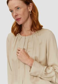 Beige zijden blouse met geplooide voorkant, ronde halslijn en lange mouwen. De gladde textuur en knoopmanchetten benadrukken het ontwerp.