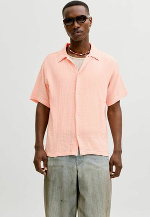 Homme portant des lunettes de soleil noires, une chemise corail à manches courtes avec boutons, un collier de perles et un pantalon ample gris clair sur un fond uni.