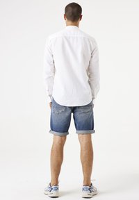 Camisa blanca de manga larga con cuello puntiagudo, combinada con pantalones cortos de mezclilla enrollados. El modelo lleva zapatillas deportivas ligeras con acentos azules.
