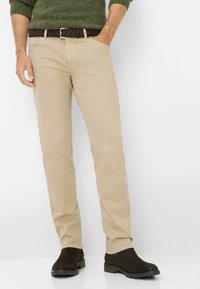 Chinos beige con taglio dritto, abbinati a una cintura marrone scuro e stivali alla caviglia marroni. Il materiale appare morbido con texture minima.
