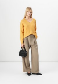Donna in maglione senape a scollo a V e pantaloni beige a gamba larga tiene una borsa nera, indossando scarpe piatte nere, in piedi di fronte a uno sfondo bianco uniforme.