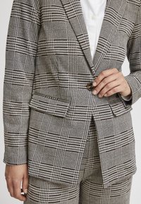 Rutigt kavaj i svart, vitt och beige tyg. Har strukturerade slag, framfickor och en enkel knappstängning.