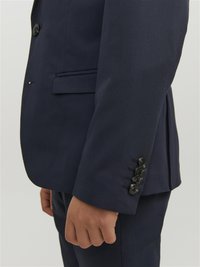 La giacca del completo navy presenta una vestibilità slim, un tessuto testurizzato, rever stretto e quattro bottoni neri sui polsini. Tasca laterale visibile.