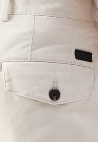 Pantalons en coton beige avec une poche arrière arborant une étiquette logo noire et un bouton sombre, des coutures détaillées et une texture lisse.