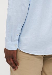 Chemise à manches longues bleu clair avec un détail de poignet boutonné, présentant une texture lisse et un ourlet arrondi. Associée à un pantalon beige clair.