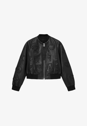 Veste aviateur noire en cuir avec des patchs brodés, y compris des chiffres et des symboles. Comprend une fermeture éclair à l'avant et des poignets côtelés.