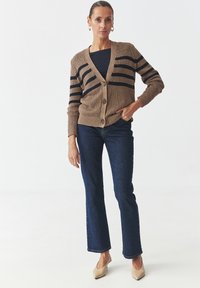 Brown knit cardigan with dark stripes, button closure, and ribbed cuffs. Paired with dark blue flared jeans and beige heels.