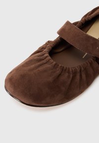 Zapato estilo Mary Jane en ante marrón con punta redondeada, detalle de fruncido alrededor de la abertura y suela plana. Cuenta con una correa ajustable.