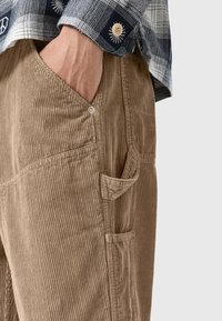 Ruskeat corduroy-haalarit, joissa on pystyribbaus, sivutaskut ja nappikoristelu. Yhdistetty sinivalkoruutuiseen flanellipaitaan.