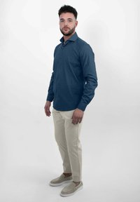 Dunkelblaue Langarm-Hemd mit Knopfleiste, beige schmal geschnittene Hose und helle Schuhe. Model steht in Pose vor einem schlichten weißen Hintergrund.