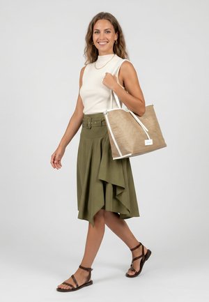Donna che cammina, indossa un top bianco senza maniche, una gonna asimmetrica verde oliva, sandali marroni, porta una grande borsa beige a tracolla.