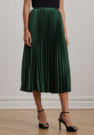 Femme portant une jupe midi plissée vert foncé associée à des talons ouverts noirs à brides à la cheville, debout sur un sol en bois.