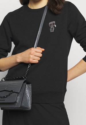 Person trägt schwarzen Pullover mit kleinem Charakter-Design und hält eine strukturierte graue Handtasche mit Kettenriemen über der Schulter.