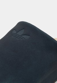 Tmavomodré semišové nazúvacie topánky so zvýrazneným detailom loga na vrchu. Sól sa javí ako svetlý, čo kontrastuje s materiálom.