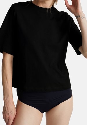 Person trägt ein locker sitzendes schwarzes T-Shirt mit kurzen Ärmeln und schwarze Unterwäsche, mit einem kleinen Tattoo sichtbar am erhobenen rechten Unterarm.
