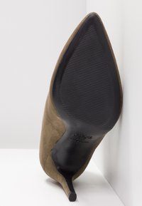 Chaussure à talon haut en daim marron à bout pointu, inclinée pour montrer la semelle noire texturée et le talon fin sur fond blanc.