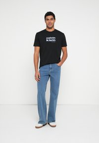 Czarny bawełniany T-shirt z nadrukiem "Calvin Klein" i "New York City" w kolorze białym i jasnoniebieskim, zestawiony z niebieskimi rozszerzanymi dżinsami i białymi butami.