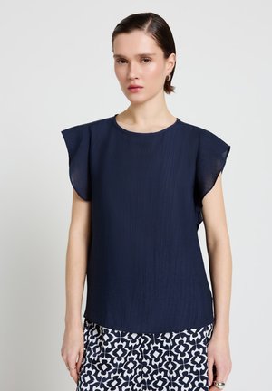 Frau trägt eine marineblaue, ärmellose Bluse mit flatternden Ärmeln und gemusterte schwarz-weiße Hose vor einem einfarbigen Hintergrund.