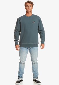 Sudadera de cuello redondo en color verde azulado con puños acanalados, de corte informal, presenta un parche de logo negro en el pecho y estampado blanco en las mangas, combinada con jeans claros.