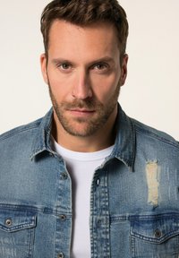 Chaqueta de mezclilla azul claro con dos bolsillos en el pecho, cierre de botones y un parche desgastado en el hombro derecho, llevada sobre una camiseta blanca.