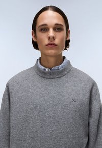 Maglione grigio lavorato a maglia con un alto collo, caratterizzato da un colletto a coste e un piccolo logo ricamato sul petto. Colletto a righe in vista.