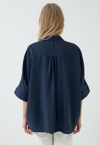 Marineblauwe, kortemouw shirt met een relaxte pasvorm, plooi detail op de rug en een gladde textuur. Gemaakt van een lichte stof.