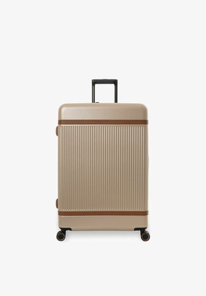 Valigia rigida beige con scanalature verticali, rifiniture marroni, manico nero retrattile e quattro ruote spinner nere su sfondo bianco.