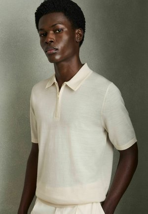 Polo shirt beige a maniche corte con zip, caratterizzato da una trama morbida e un collo classico. Indossato da un modello con uno sfondo neutro.