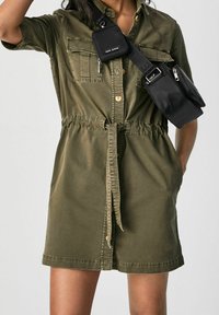 Olivgrön kortärmad skjortklänning i bomull, med två bröstfickor, en insvängd midja och ett matchande bälte i samma tyg. Svart axelremsväska i tillägg.