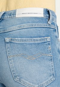 Jeans en denim bleu clair avec une texture légèrement usée. Présente une poche arrière avec un motif de couture tissé distinctif et une étiquette de marque.
