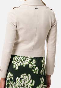 Beige cropped jakke med en glat tekstur, der har en klassisk krave, skulderstropper og en subtil sømdetalje på ryggen.