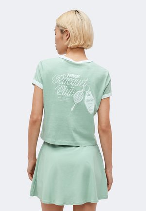 Žena s krátkymi blond vlasmi, ktorá nosí mätovo zelené tričko Nike Racquet Club a zodpovedajúcu sukňu, zobrazená zozadu.