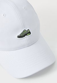 Casquette de baseball en coton blanc avec un design de baskets brodé en noir et vert fluo à l'avant. Comprend des trous de ventilation cousus et une visière incurvée.