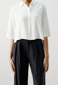 Camicia bianca corta con colletto, maniche corte e finitura testurizzata, abbinata a pantaloni scuri e morbidi.