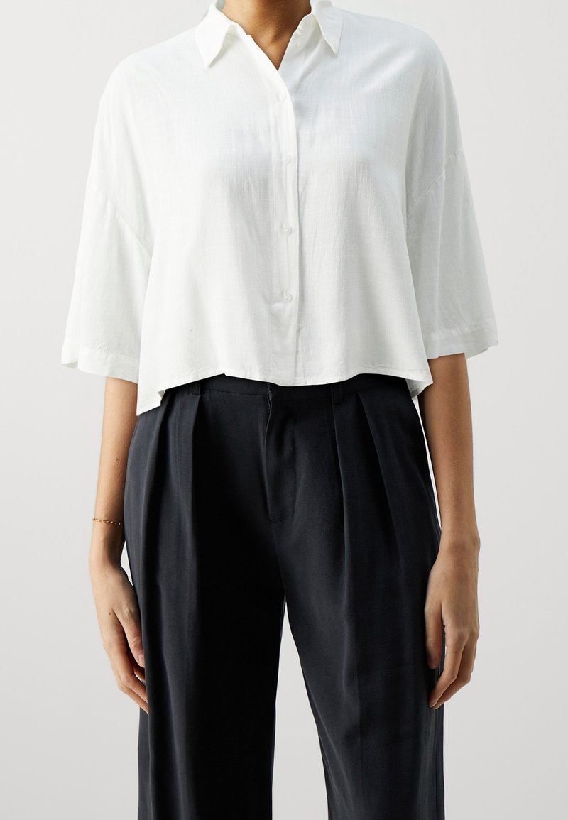 Camicia bianca corta con colletto, maniche corte e finitura testurizzata, abbinata a pantaloni scuri e morbidi.