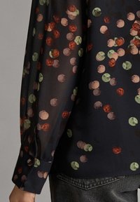 Genomskinlig svart blus med flerfärgade cirkulära prickar i rött, grönt och persika. Långa ärmar med knäppning vid handleden och en mjuk fall.