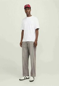 Man met een rode pet, een wit oversized t-shirt, losse grijze spijkerbroek en witte sneakers tegen een effen achtergrond.