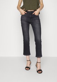 Jean en denim foncé coupe slim et longueur raccourcie, avec un ourlet brut. Associé à des sandales noires à lanières et un haut côtelé vert olive.