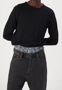 Persona vestida con una camisa de manga larga negra metida dentro de unos vaqueros oscuros, con una cinturilla de cuadros azules visible debajo.