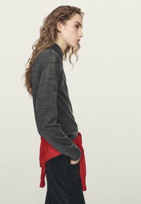 Gray long-sleeve shirt made of textured fabric, featuring a high neckline. A red sweater is tied around the waist, contrasting with dark pants.