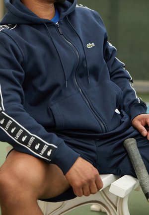 Mann sitzt auf einer weißen Bank, trägt einen dunkelblauen Lacoste-Hoodie und Shorts und hält einen Tennisschläger mit abgenutztem Griff.