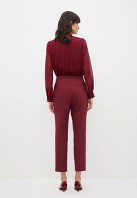 Blusa bordeaux con plissè verticali e morbida texture, abbinata a pantaloni bordeaux su misura. Entrambi hanno un design elegante e aderente.