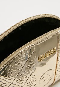 Guldmetallic pose med quiltet mønster, med lynlås og "GUESS" logo i gyldne bogstaver på fronten. Sort indvendig foring.