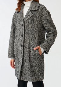 Kvinde iført en sort-hvid herringbone mønstret frakke med stor krave og sorte knapper, hånd i lommen, over hvid turtleneck og sorte bukser.